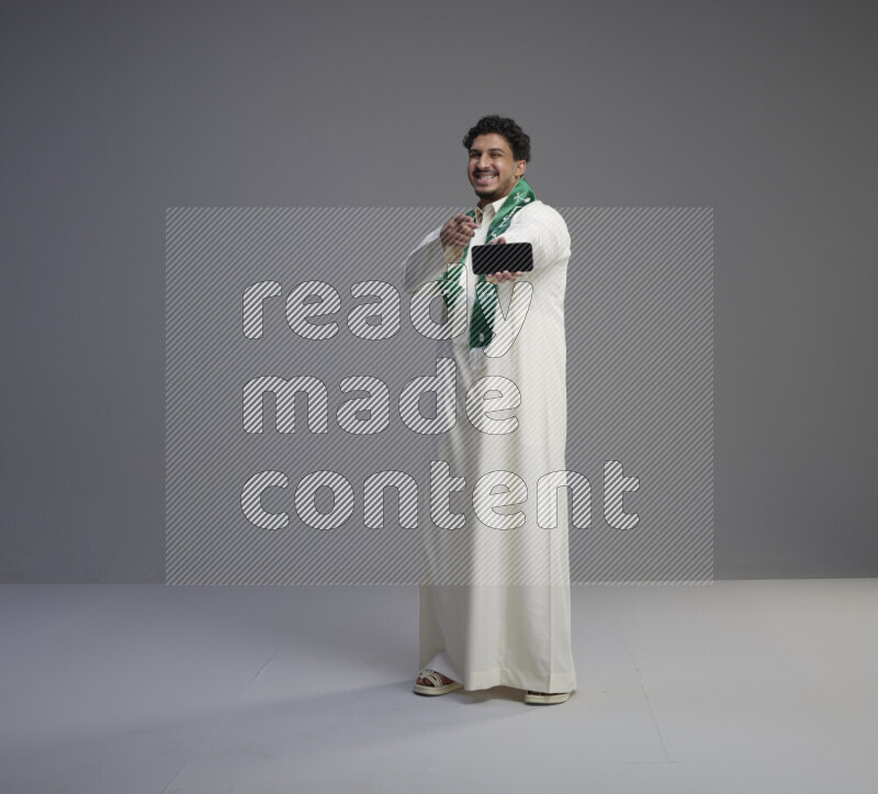 A Saudi man standing wearing thob and Saudi flag scarf showing phone to camera on gray background