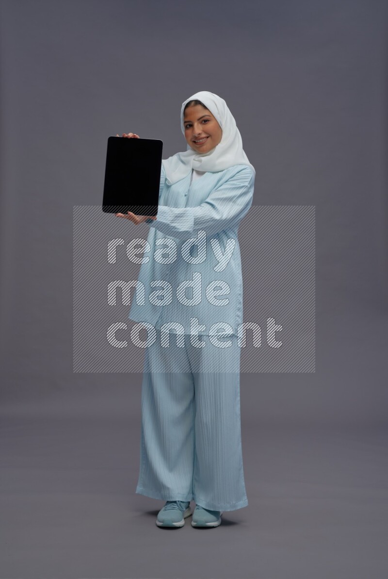 Saudi woman wearing hijab clothes standing showing tablet to camera on gray background