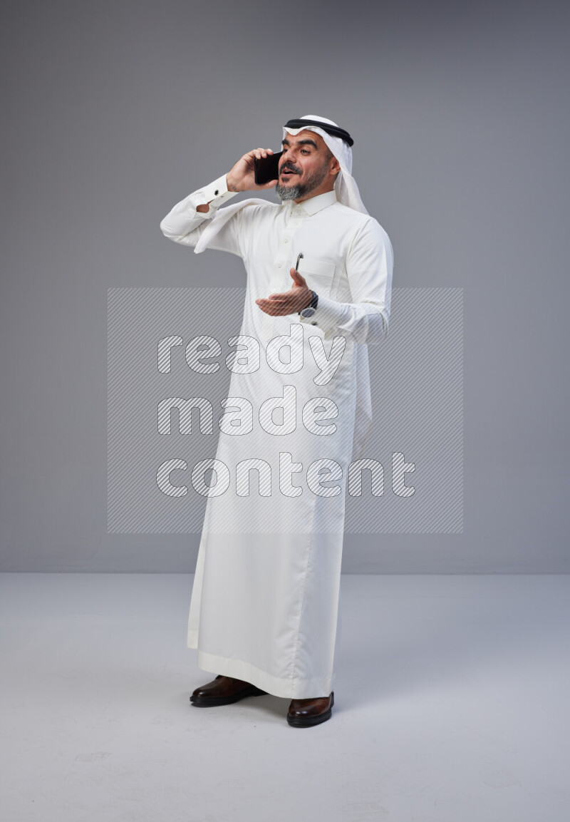 Saudi man Wearing Thob and white Shomag standing talking on phone on Gray background