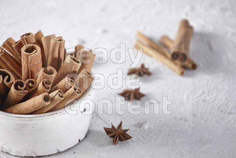 White bowl full of cinnamon sticks surrounded by star anis on a textured white background in different angles