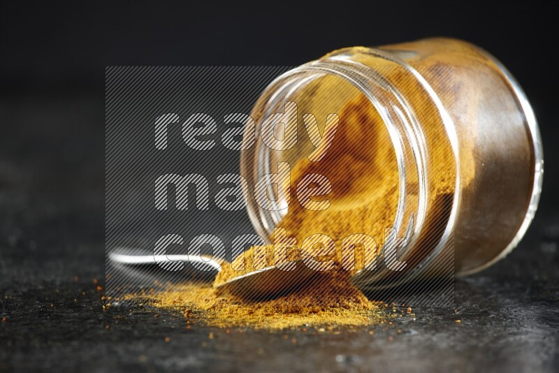 A flipped glass jar and a metal spoon full of turmeric powder and powder spilled out of it on textured black flooring
