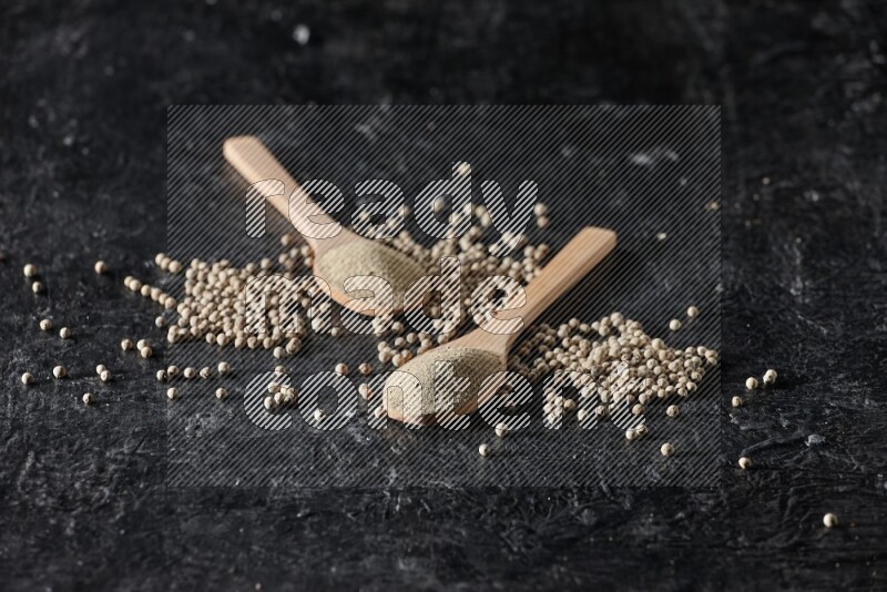 2 wooden spoons full of white pepper powder with white pepper beads on textured black flooring