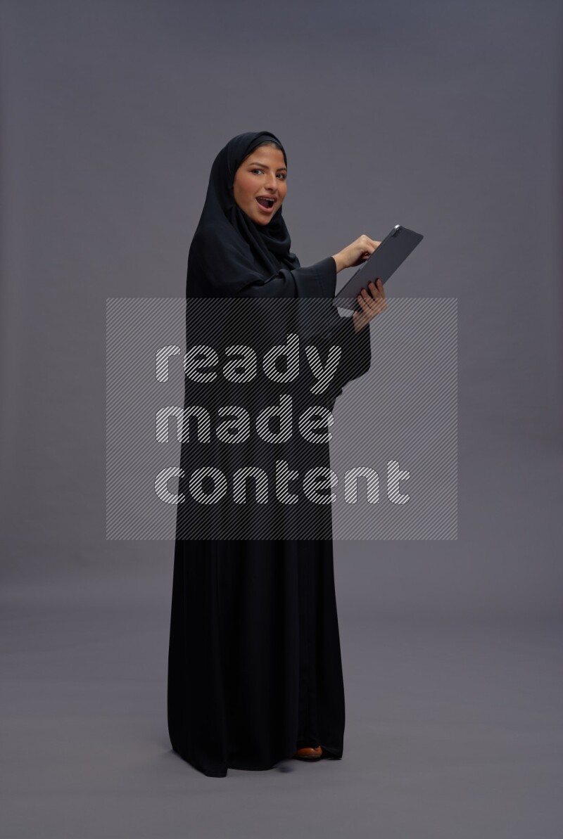 Saudi woman wearing Abaya standing working on tablet on gray background