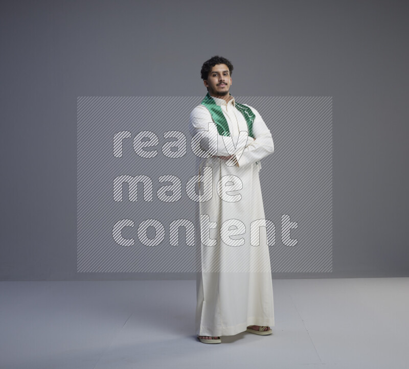 A Saudi man standing wearing thob and Saudi flag scarf on gray background