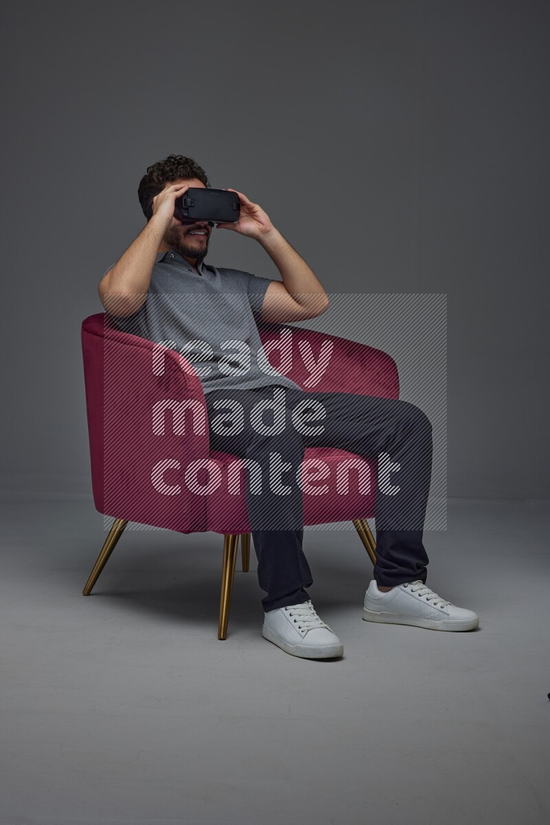 A man wearing casual and wearing VR while sitting on a burgundy chair eye level on a gray background
