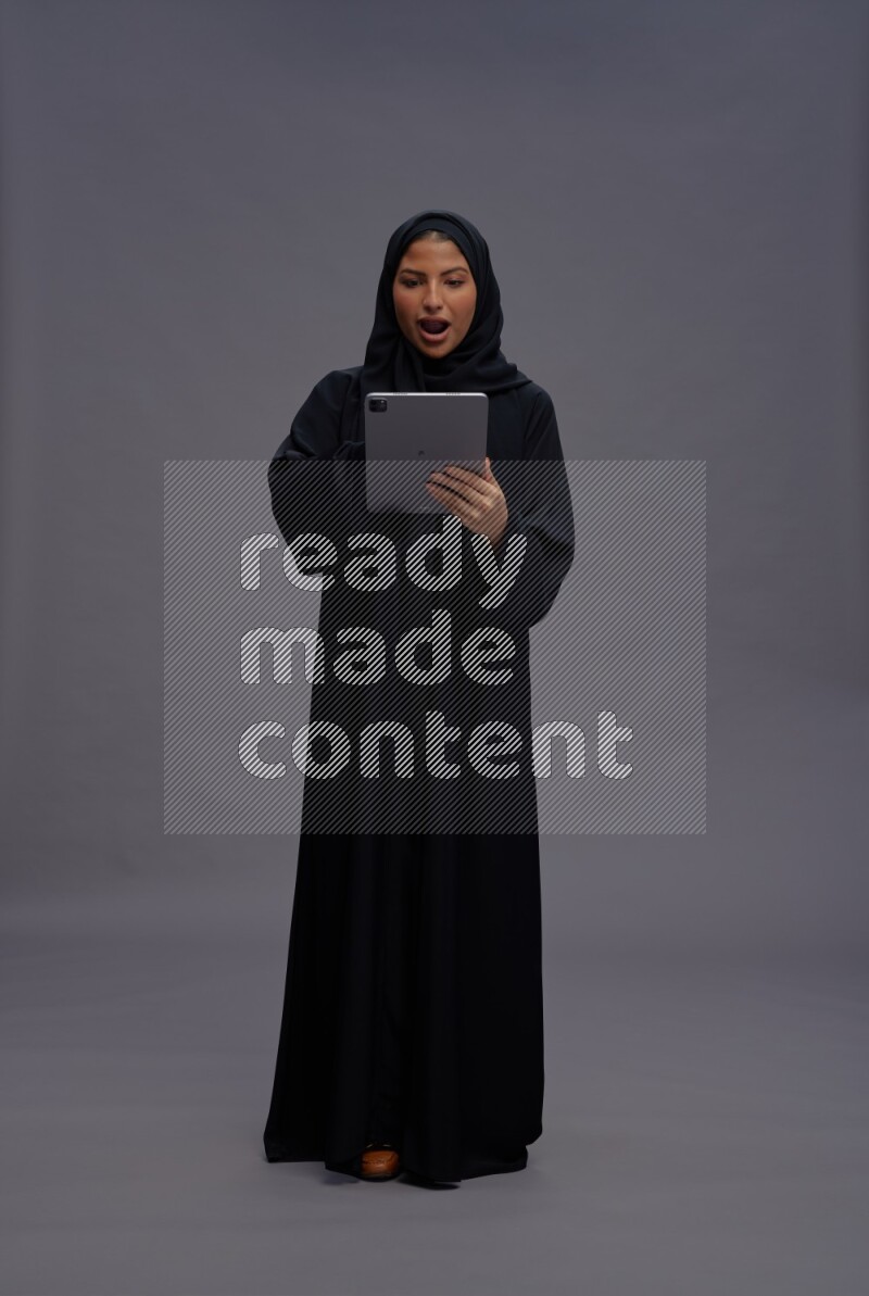Saudi woman wearing Abaya standing working on tablet on gray background