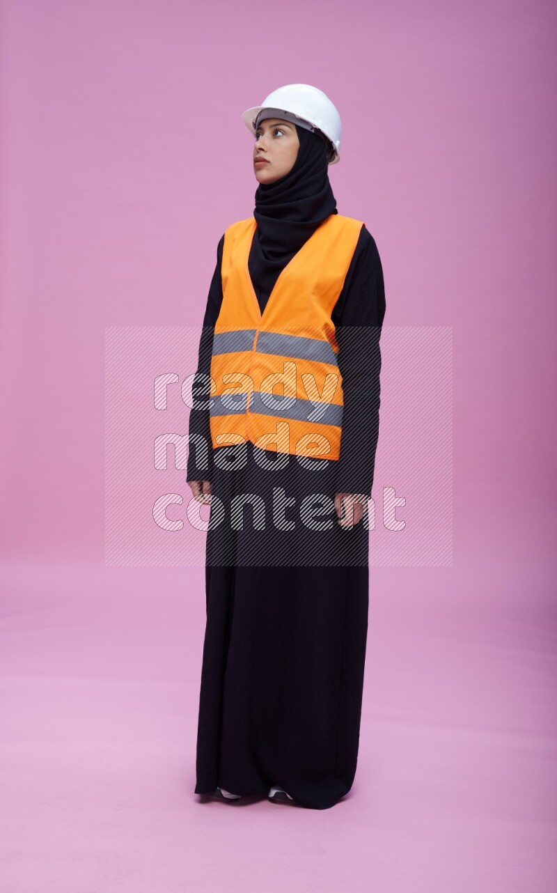 Saudi woman wearing Abaya with engineer vest and helmet standing interacting with the camera on pink background