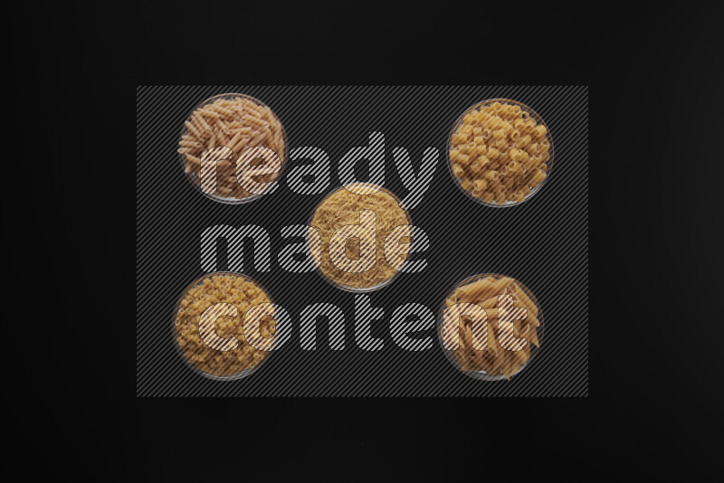 Different pasta types in 5 glass bowls on black background