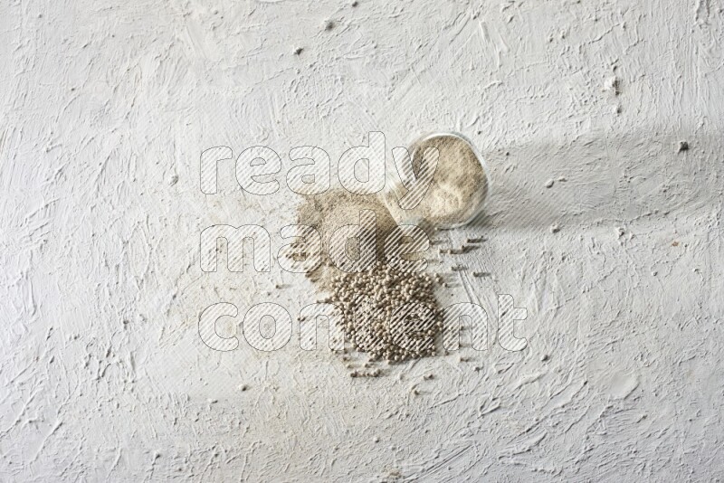 A flipped herbal glass jar full of white pepper powder with spilled powder and beads on textured white flooring