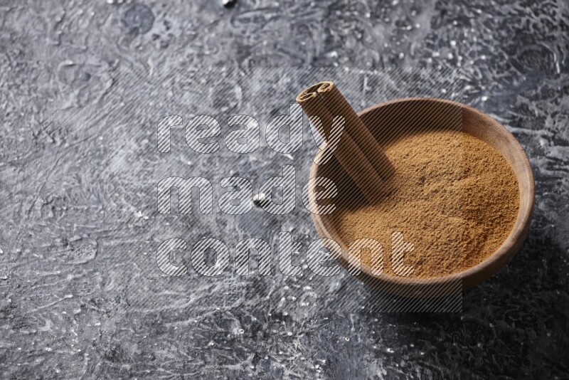 Wooden bowl full of cinnamon powder and a cinnamon stick on a textured black background