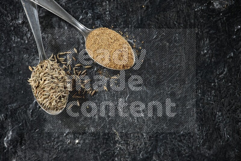 2 metal spoons full of cumin seeds and cumin powder on a textured black flooring