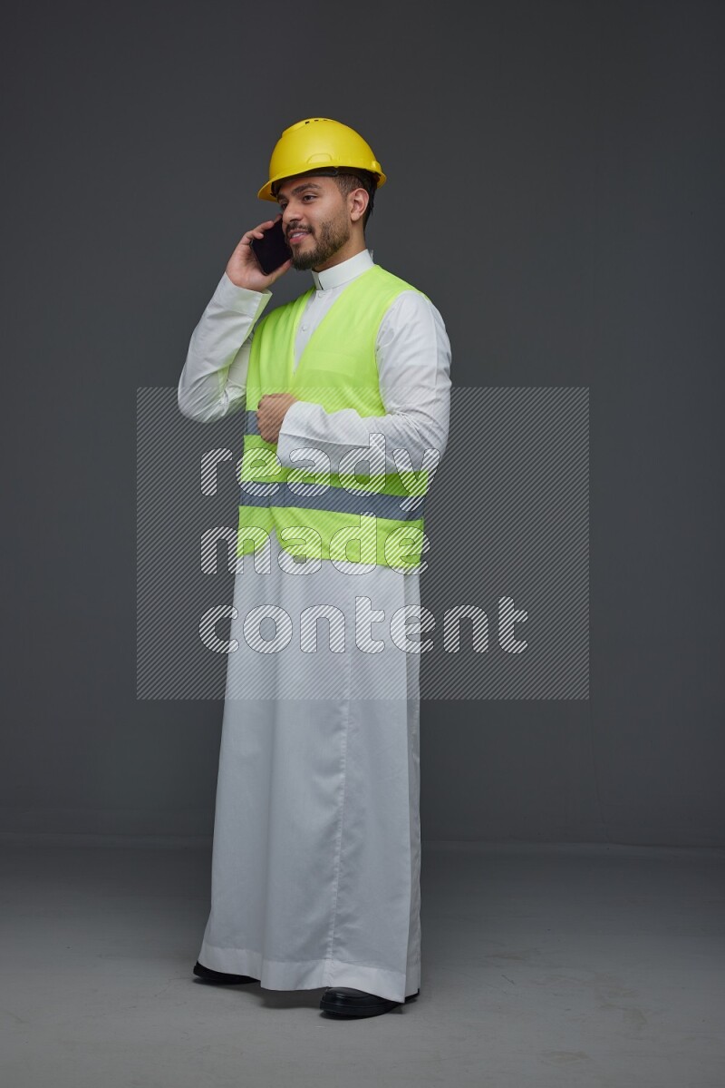 A Saudi man wearing Thobe with a yellow safety vest and white helmet standing and talking in the phone eye level on a gray background