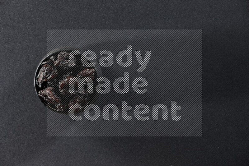A black pottery bowl full of dried plums on a black background in different angles