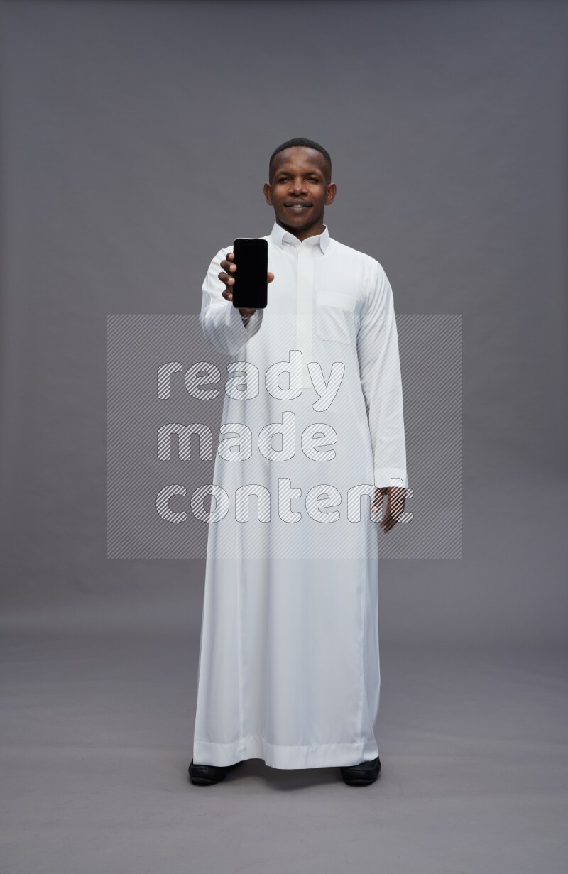 Saudi man wearing thob standing showing phone to camera on gray background
