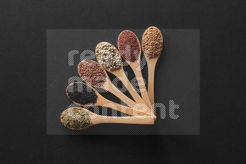 wooden spoons filled with white peppers, mustard seeds, black seeds, garden cress, cumin and flax on black flooring