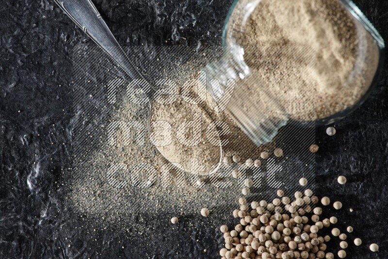 A flipped herbal glass jar and metal spoon full of white pepper powder with spilled powder and pepper beads on textured black flooring