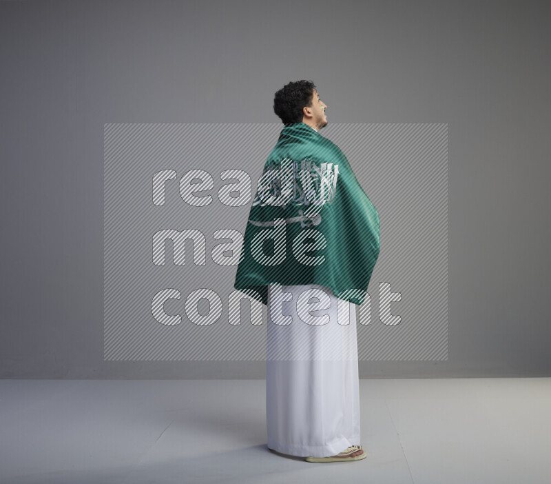 A Saudi man standing wearing thob with face painting wrapping big Saudi flag on gray background