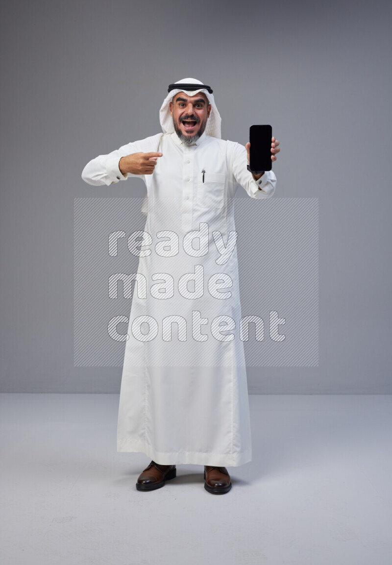 Saudi man Wearing Thob and white Shomag standing showing phone to camera on Gray background