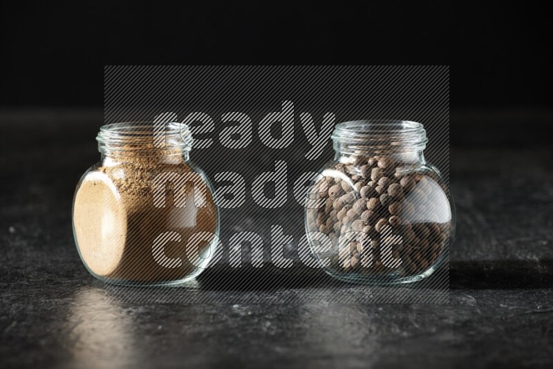 2 glass spice jars full of allspice powder and whole balls on a textured black flooring