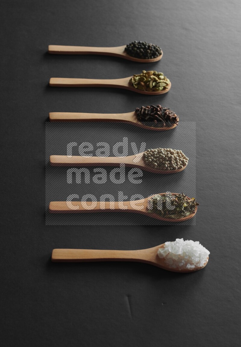 wooden spoons filled with white peppers, cloves, cardamom, salt, black peppers and basil on black flooring
