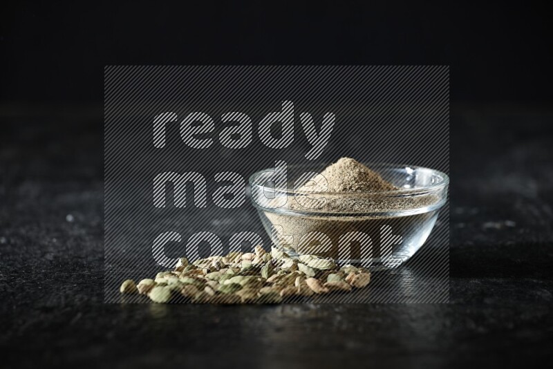A glass bowl full of cardamom powder and cardamom seeds beside it on textured black flooring