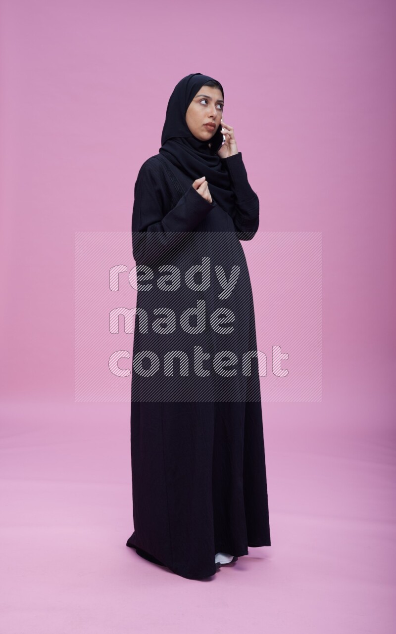 Saudi woman wearing Abaya standing talking on phone on pink background