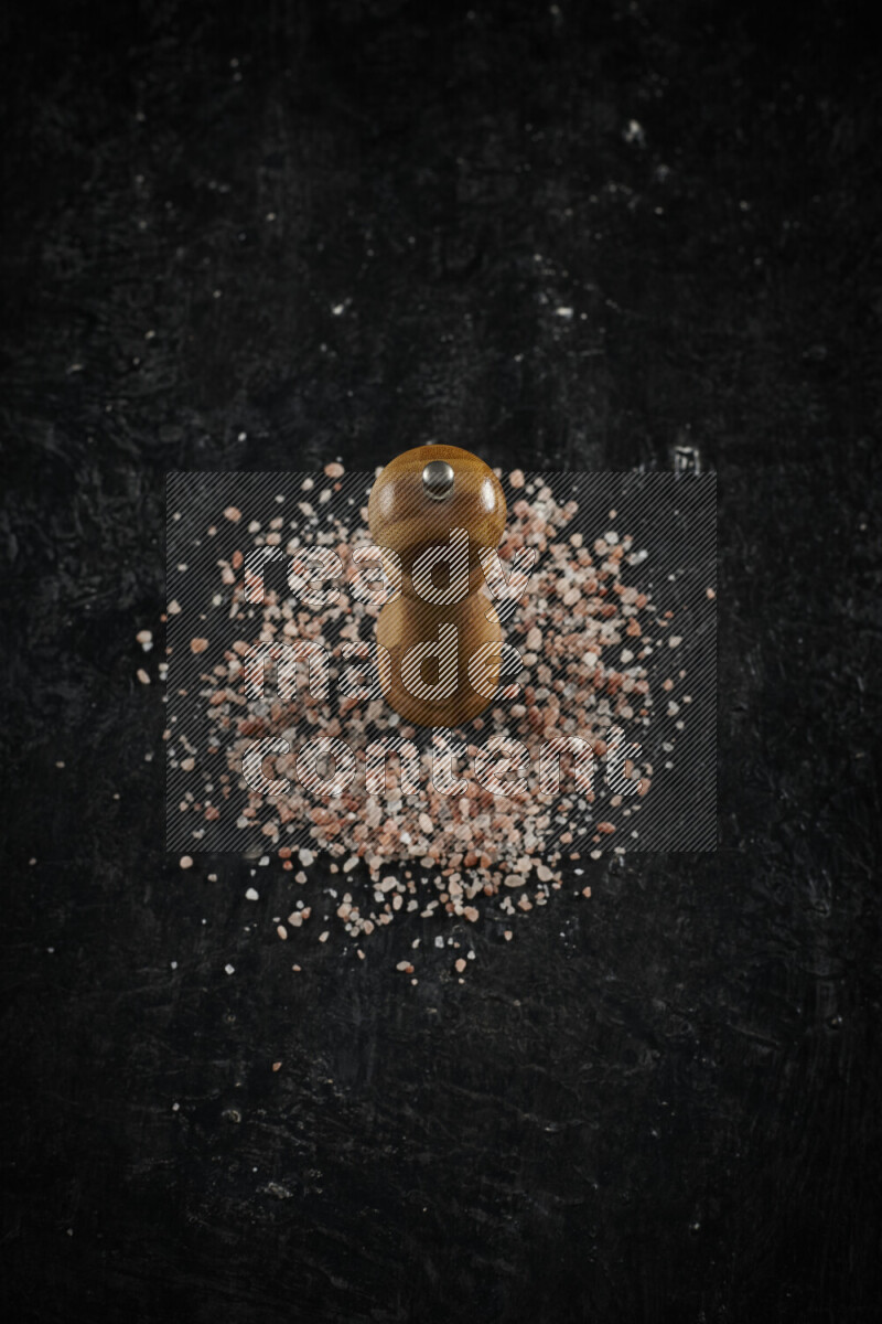 A wooden grinder standing upright and surrounded by coarse pink himalayan salt on black background
