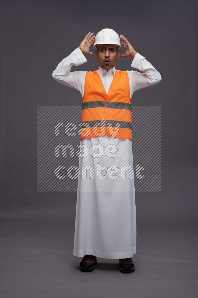 Saudi man wearing thob with engineer vest standing hands on head on gray background