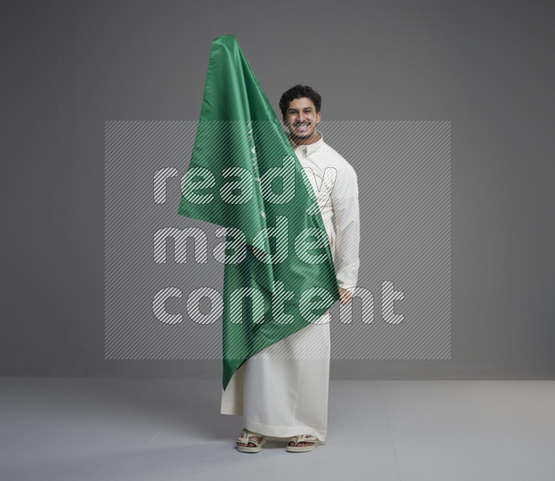 A Saudi man standing wearing thob holding big Saudi flag on gray background
