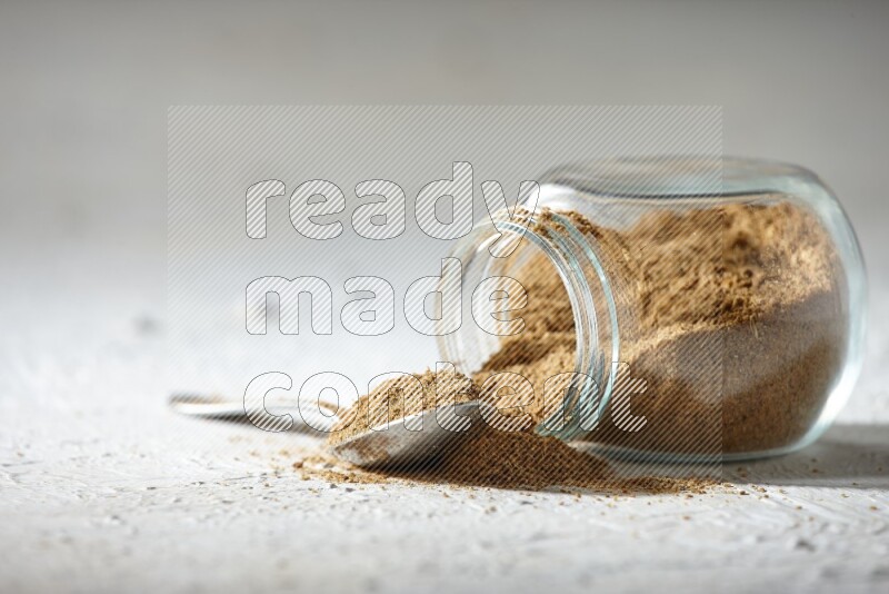 A flipped glass spice jar and a metal spoon full of cumin powder and powder spilled out on textured white flooring