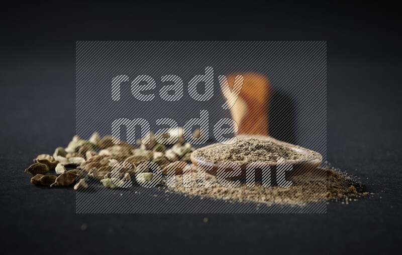 A wooden ladle full of cardamom powder and cardamom seeds beside it on black flooring