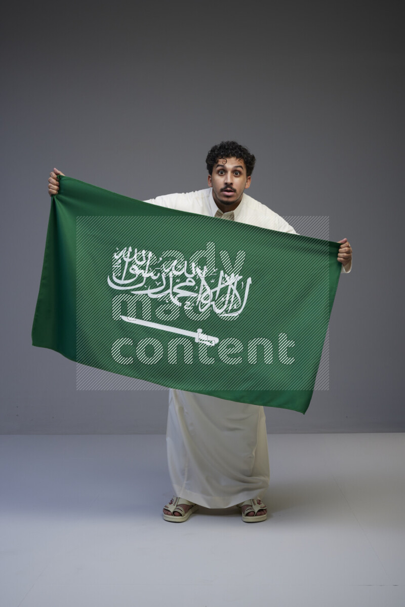 A Saudi man standing wearing thob holding big Saudi flag on gray background