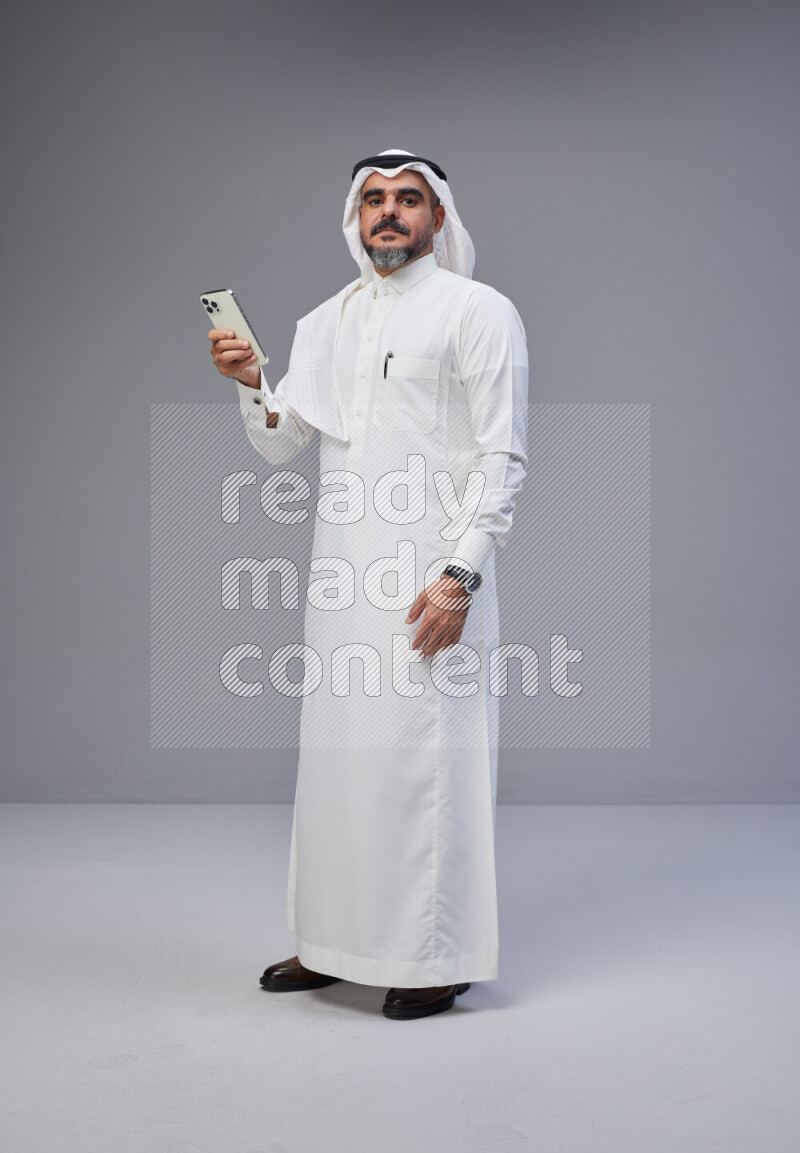 Saudi man Wearing Thob and white Shomag standing texting on phone on Gray background