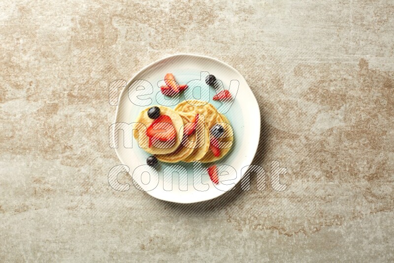 Five stacked mixed berries mini pancakes in a bicolor plate on beige background