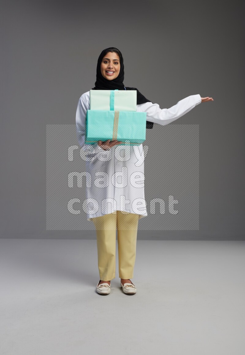 Saudi woman wearing lab coat with stethoscope standing holding gift box on Gray background