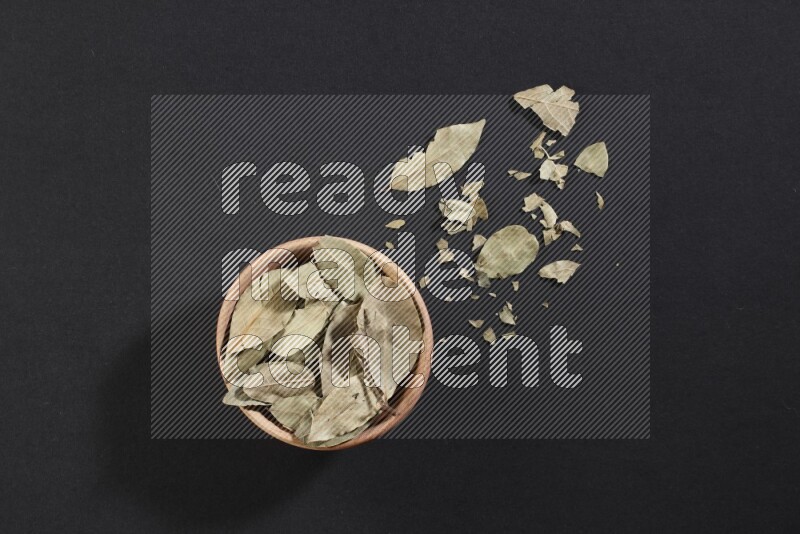 A wooden bowl filled with dried bay leaves on black flooring