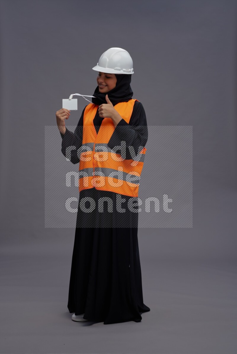 Saudi woman wearing Abaya with engineer vest with neck strap employee badge standing interacting with the camera on gray background