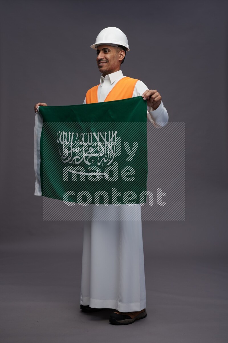 Saudi man wearing thob with engineer vest standing holding Saudi flag on gray background