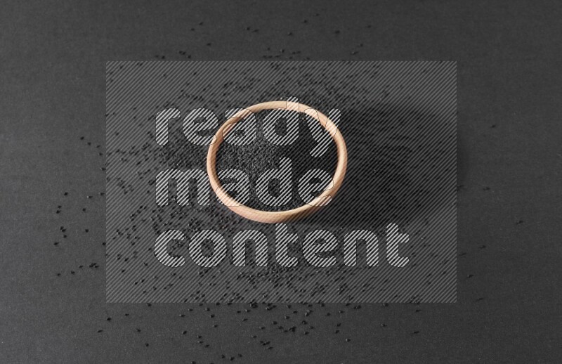 A wooden bowl full of black seeds surrounded by seeds on a black flooring