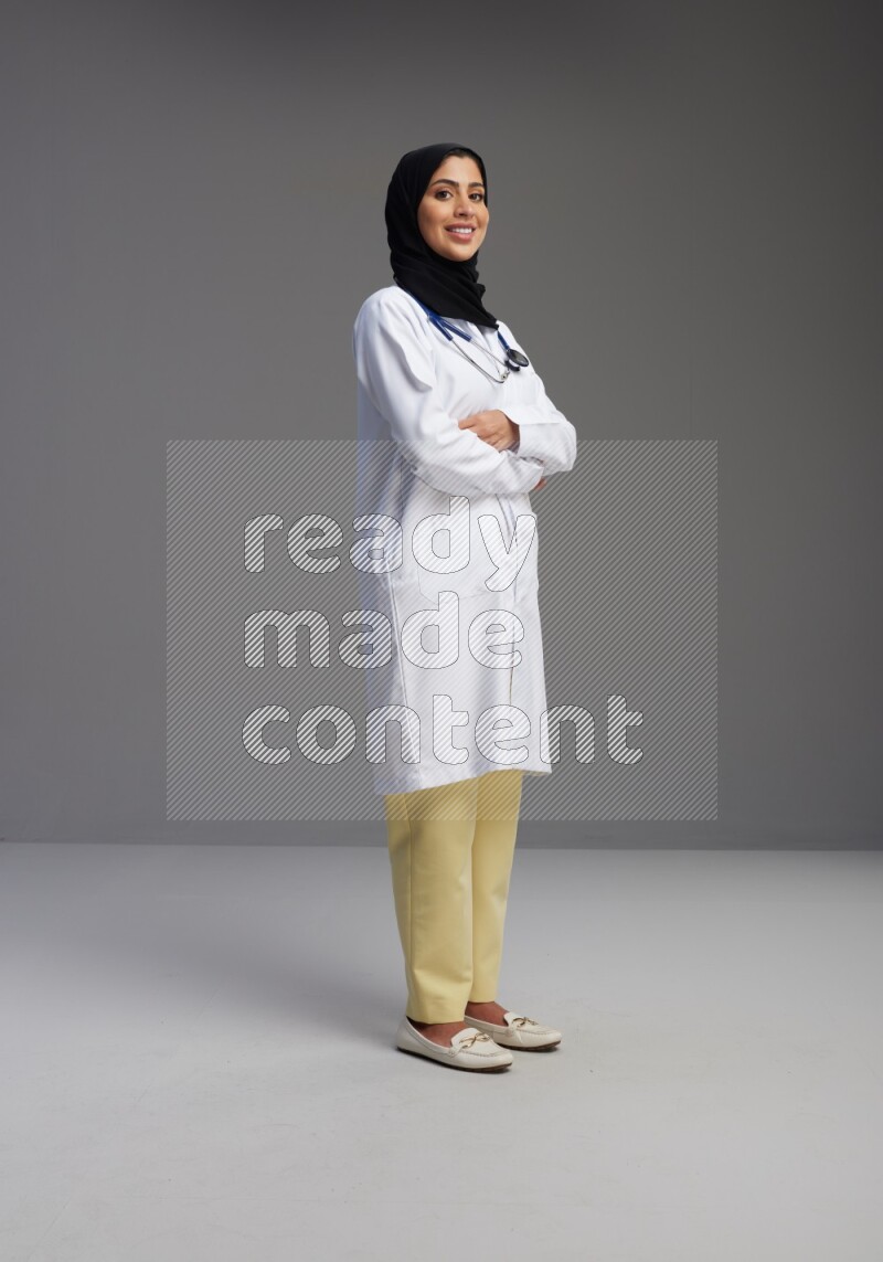 Saudi woman wearing lab coat with stethoscope standing with crossed arms on Gray background