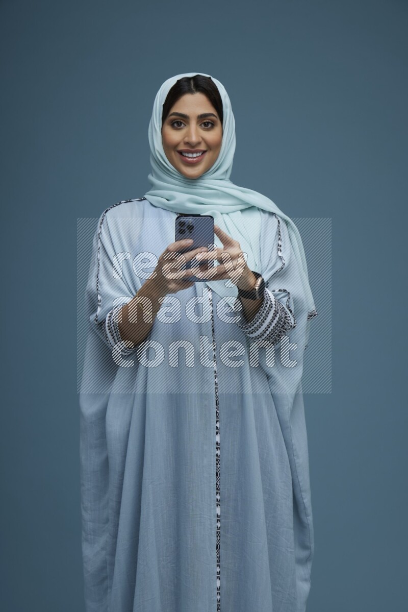 A Saudi woman posing with her phone in a blue background wearing a blue Abaya with hijab
