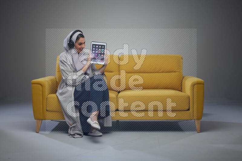 A Saudi female wearing a light gray Abaya and head scarf sitting on a yellow sofa and showing her tablet's screen while wearing headphones eye level on a grey background