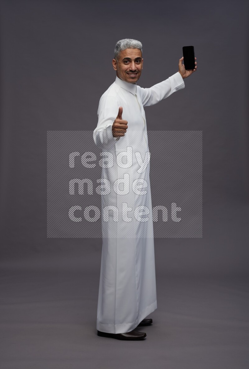 Saudi man wearing thob standing taking selfie on gray background