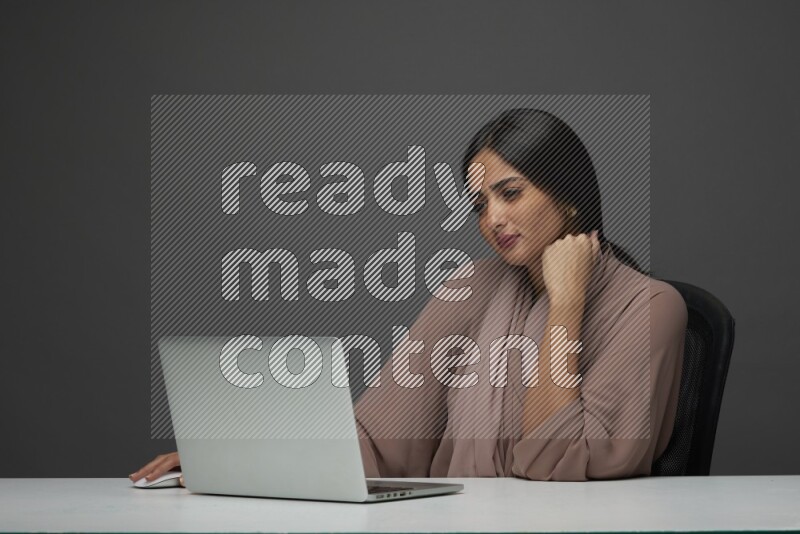 A Saudi woman Sitting on her desk on a Gray Background wearing Brown Abaya