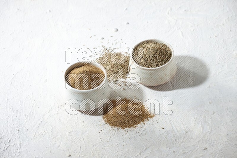 2 beige bowls full of cumin seeds and powder with spilled powder and seeds on textured white flooring