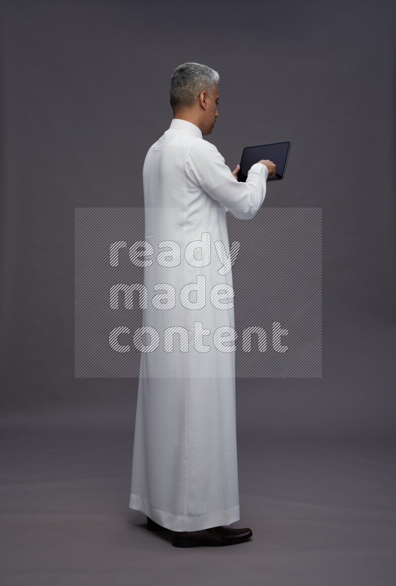 Saudi man wearing thob standing working on tablet on gray background