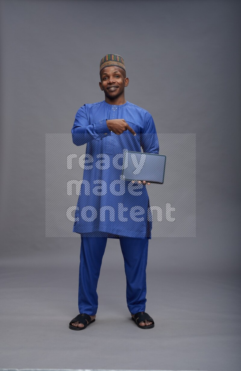 Man wearing Nigerian outfit standing showing tablet to camera on gray background