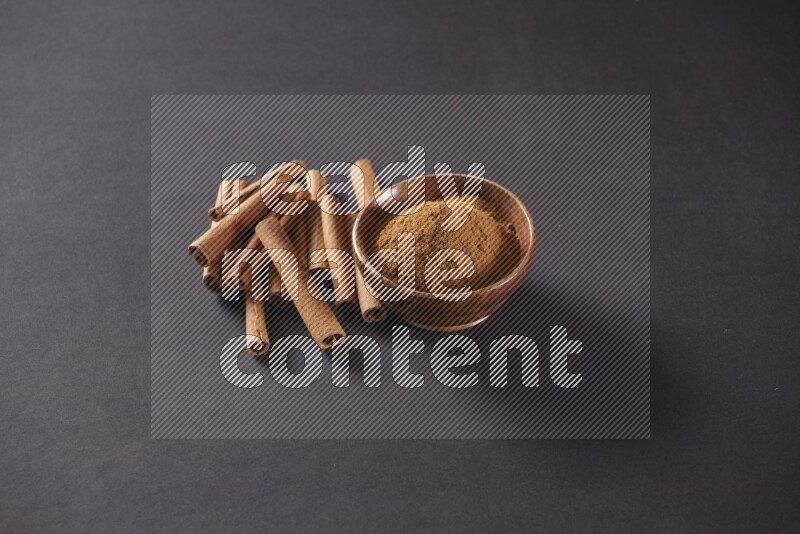 Cinnamon sticks stacked beside a wooden bowl full of cinnamon powder on black background