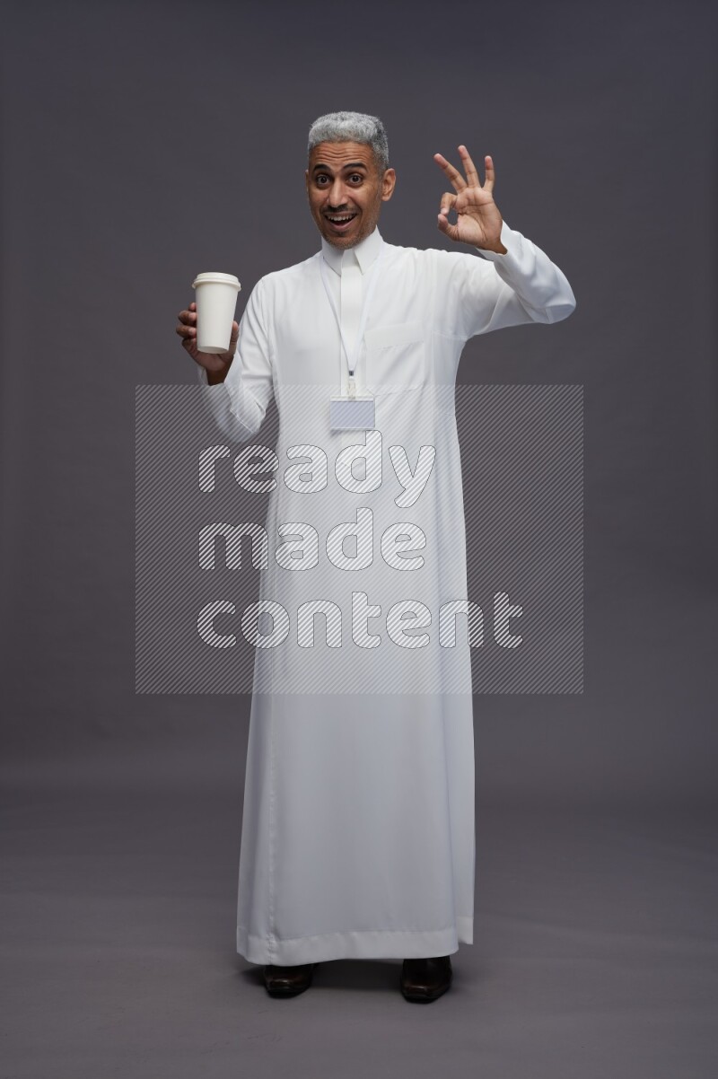 Saudi man wearing thob with neck strap employee badge standing holding paper cup on gray background