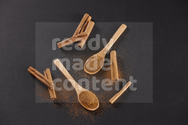 Cinnamon powder in two wooden spoons with cinnamon sticks on black background